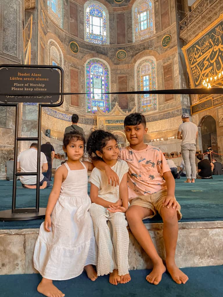 A boy and two little girls sitting with a mosque behind them