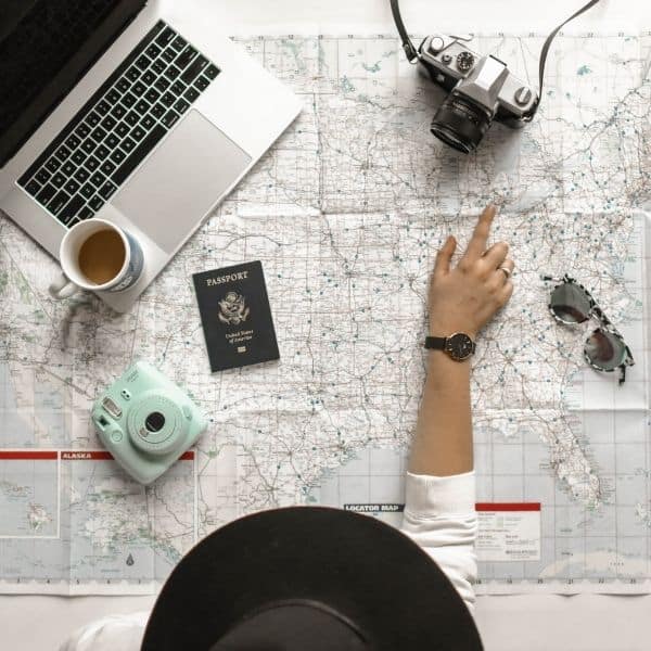 A flatlay with macbook, camera, coffee, passport and camera with a woman pointing at a map